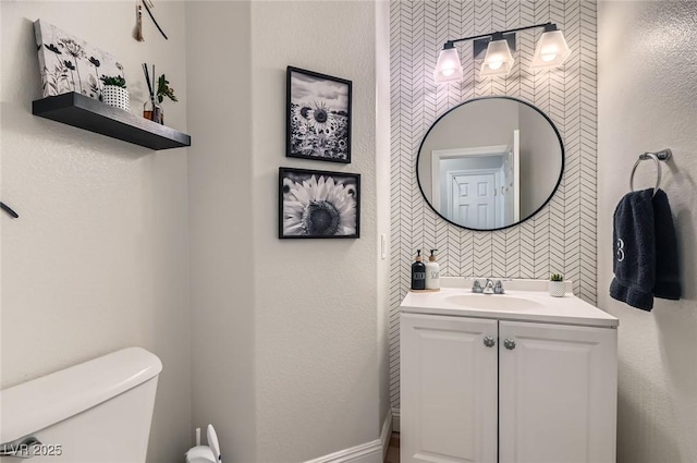 bathroom with toilet, wallpapered walls, baseboards, and vanity