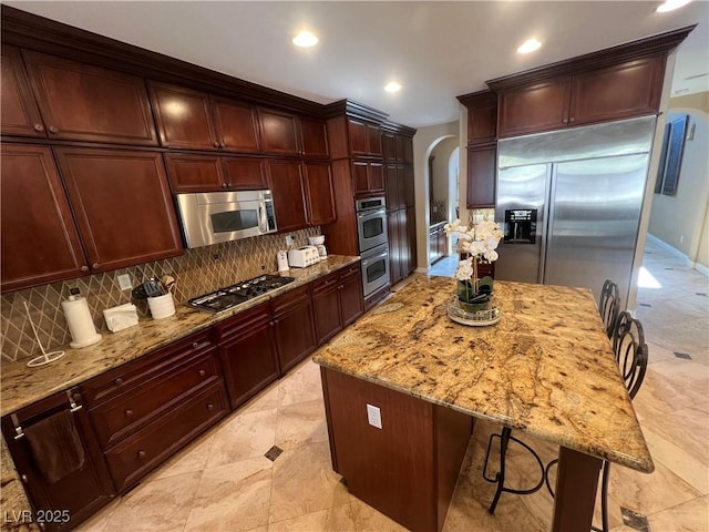 kitchen with backsplash, a kitchen bar, appliances with stainless steel finishes, arched walkways, and light stone counters