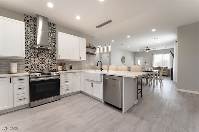 kitchen featuring appliances with stainless steel finishes, a sink, wall chimney exhaust hood, a peninsula, and light wood-type flooring