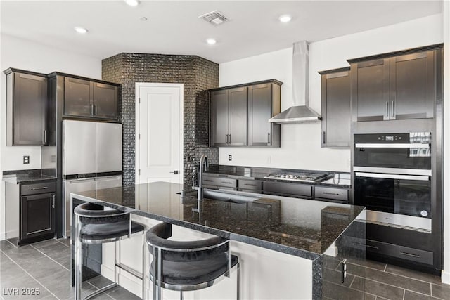 kitchen featuring visible vents, a sink, dark stone countertops, stainless steel appliances, and wall chimney exhaust hood