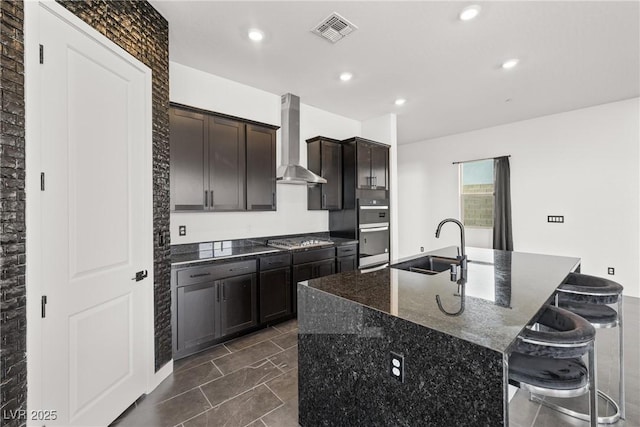 kitchen with a sink, wall chimney exhaust hood, stainless steel appliances, visible vents, and a center island with sink