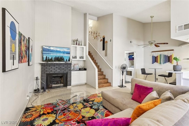 living area with a high ceiling, ceiling fan, a brick fireplace, visible vents, and stairway