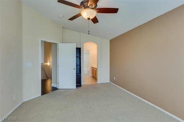 unfurnished bedroom with baseboards, ensuite bathroom, ceiling fan, light colored carpet, and arched walkways