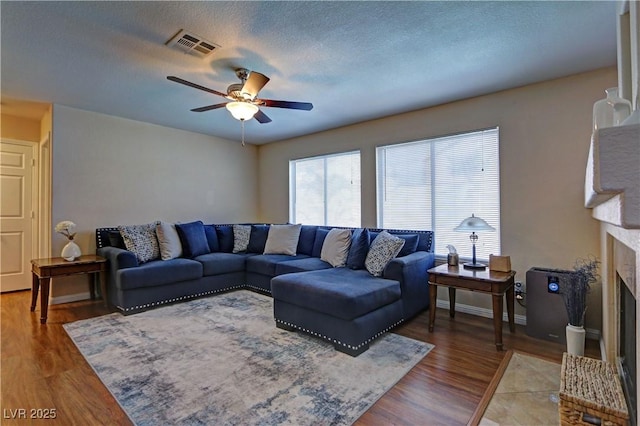 living room featuring ceiling fan, a fireplace with raised hearth, a textured ceiling, light wood-type flooring, and visible vents