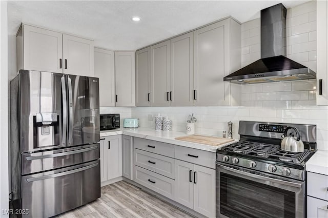 kitchen with wall chimney range hood, gray cabinets, stainless steel appliances, light wood-type flooring, and backsplash