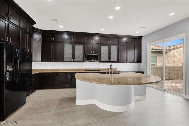 kitchen featuring light stone countertops, black fridge, a sink, a kitchen island with sink, and stainless steel microwave