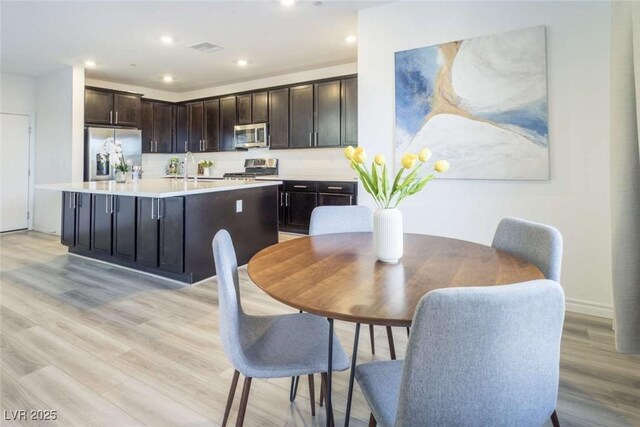 kitchen with dark brown cabinets, light wood finished floors, light countertops, stainless steel appliances, and a center island with sink