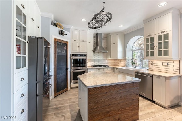kitchen with wall chimney range hood, appliances with stainless steel finishes, a sink, white cabinets, and ornamental molding