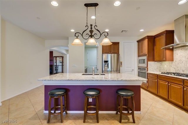 kitchen featuring wall chimney range hood, appliances with stainless steel finishes, a sink, a kitchen island with sink, and arched walkways