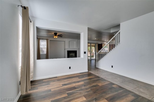 unfurnished living room featuring a ceiling fan, a fireplace, wood finished floors, baseboards, and stairs