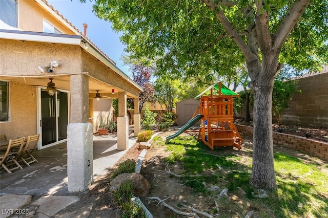 view of jungle gym featuring an outdoor structure, a patio, a fenced backyard, a storage unit, and a ceiling fan