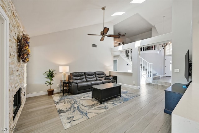 living area with stairs, a stone fireplace, high vaulted ceiling, light wood-type flooring, and a ceiling fan
