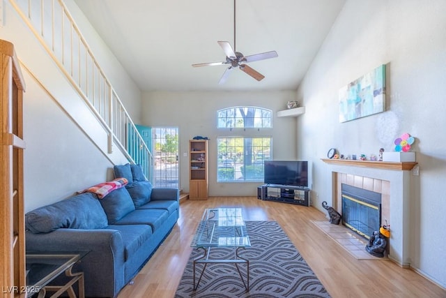 living room with a ceiling fan, a tiled fireplace, wood finished floors, stairway, and high vaulted ceiling