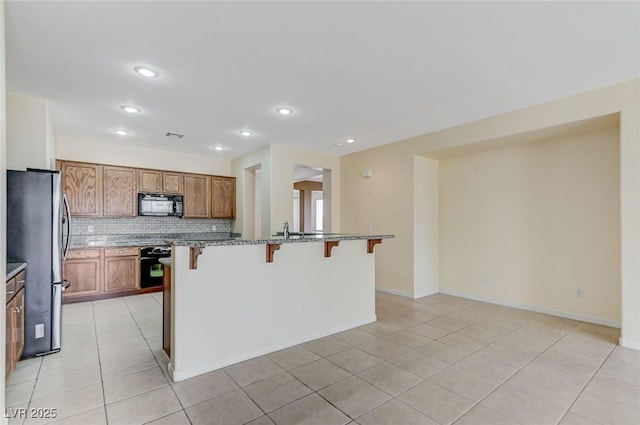 kitchen featuring black microwave, a breakfast bar, freestanding refrigerator, decorative backsplash, and light tile patterned floors