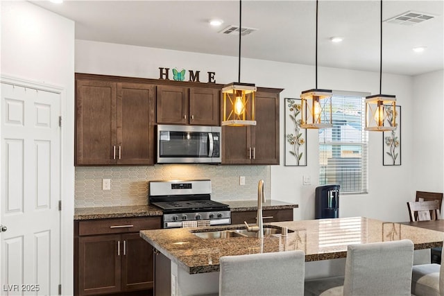 kitchen with stainless steel appliances, a sink, decorative backsplash, an island with sink, and stone counters