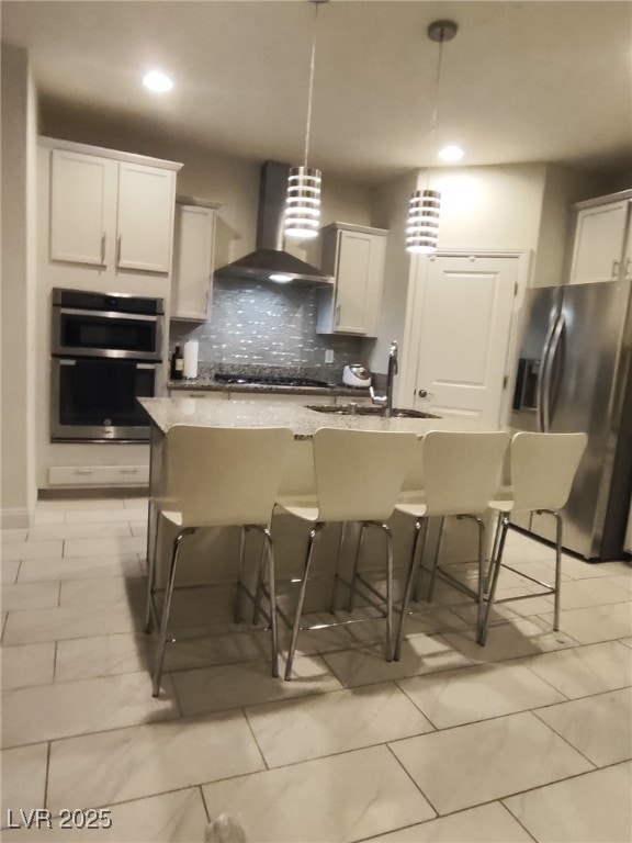 kitchen with wall chimney range hood, backsplash, a kitchen bar, appliances with stainless steel finishes, and a sink