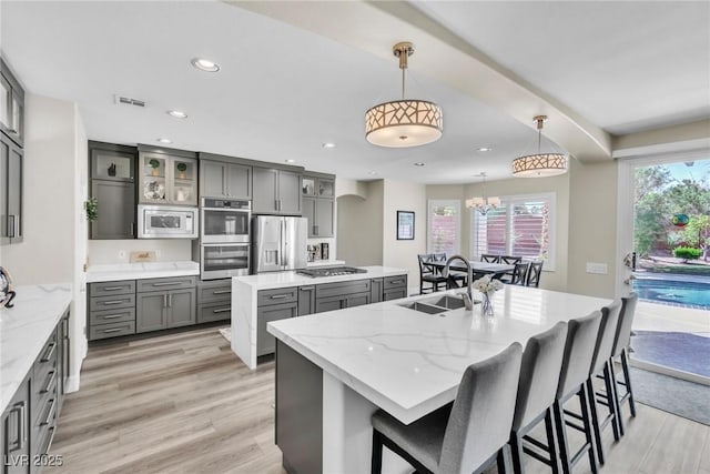kitchen with a center island with sink, gray cabinetry, a sink, glass insert cabinets, and stainless steel appliances