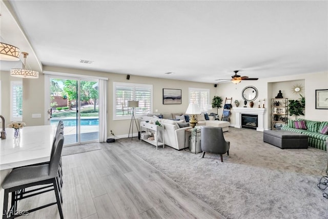 living area with visible vents, a glass covered fireplace, baseboards, a ceiling fan, and light wood-style flooring
