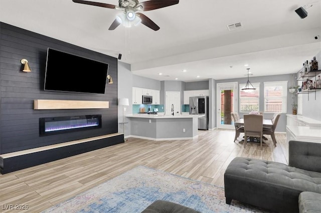 living area featuring ceiling fan, visible vents, a glass covered fireplace, and light wood-type flooring