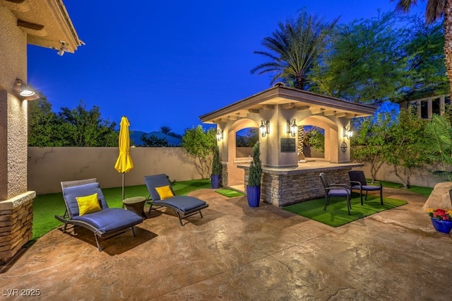 patio at twilight with a fenced backyard, outdoor dry bar, and a gazebo