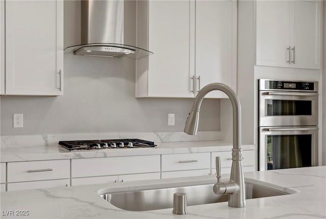 kitchen with wall chimney range hood, white cabinets, light stone counters, a sink, and appliances with stainless steel finishes