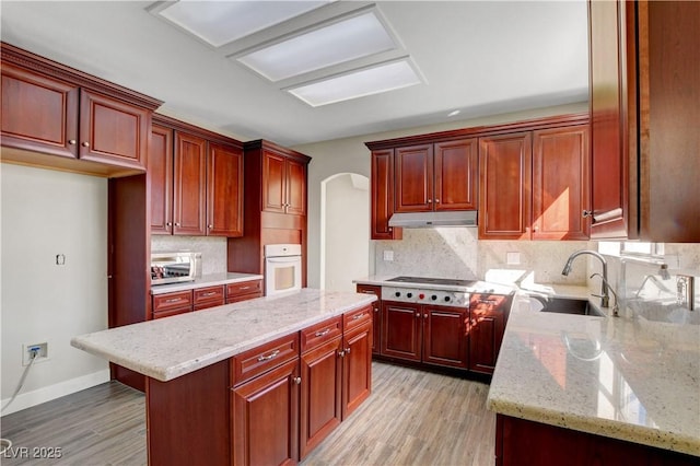 kitchen featuring oven, a sink, stainless steel cooktop, reddish brown cabinets, and under cabinet range hood