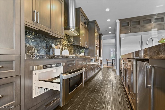 kitchen featuring wall chimney range hood, dark wood finished floors, baseboards, decorative backsplash, and glass insert cabinets