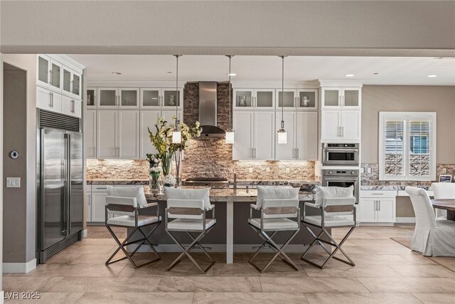 kitchen with decorative backsplash, wall chimney exhaust hood, white cabinets, light stone counters, and appliances with stainless steel finishes