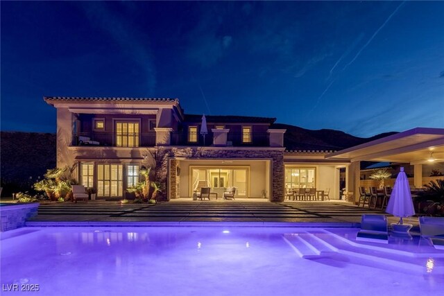 back of house at twilight featuring a balcony, a patio, an outdoor pool, stucco siding, and a tile roof