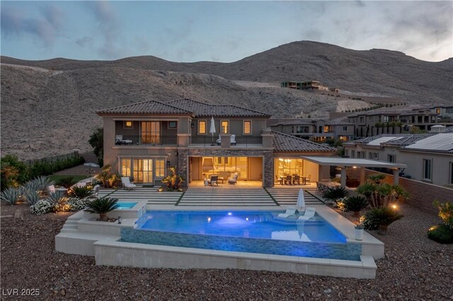 rear view of property featuring stone siding, a mountain view, an in ground hot tub, a balcony, and a patio