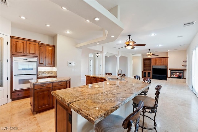 kitchen featuring brown cabinetry, double oven, ceiling fan, a spacious island, and a sink