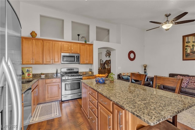 kitchen featuring light stone countertops, appliances with stainless steel finishes, a breakfast bar, arched walkways, and a ceiling fan