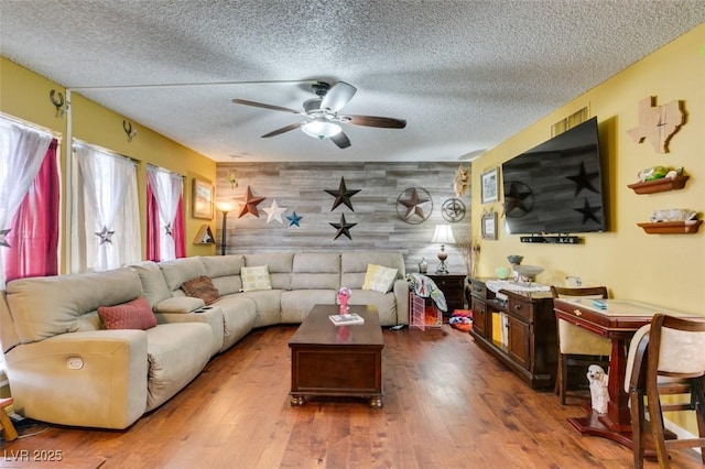 living area featuring wooden walls, a textured ceiling, an accent wall, a ceiling fan, and wood finished floors