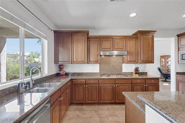 kitchen featuring brown cabinets, appliances with stainless steel finishes, light stone counters, a sink, and under cabinet range hood