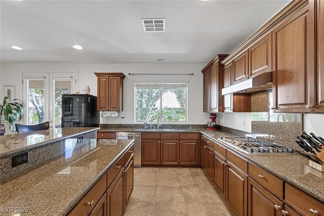 kitchen with brown cabinetry, dark stone counters, under cabinet range hood, a sink, and appliances with stainless steel finishes