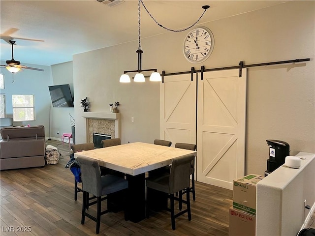 dining area featuring ceiling fan, visible vents, a barn door, a glass covered fireplace, and dark wood-type flooring