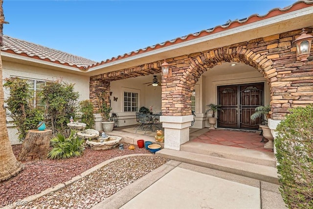 entrance to property with a ceiling fan, a tile roof, a patio, stone siding, and stucco siding