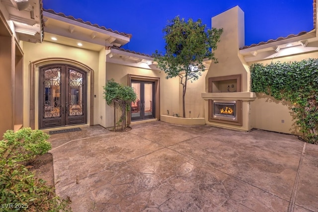 entrance to property featuring stucco siding, french doors, a patio area, a tiled roof, and a lit fireplace