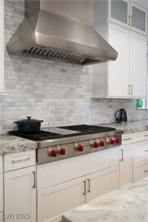 kitchen featuring wall chimney range hood, stainless steel gas stovetop, backsplash, glass insert cabinets, and white cabinetry