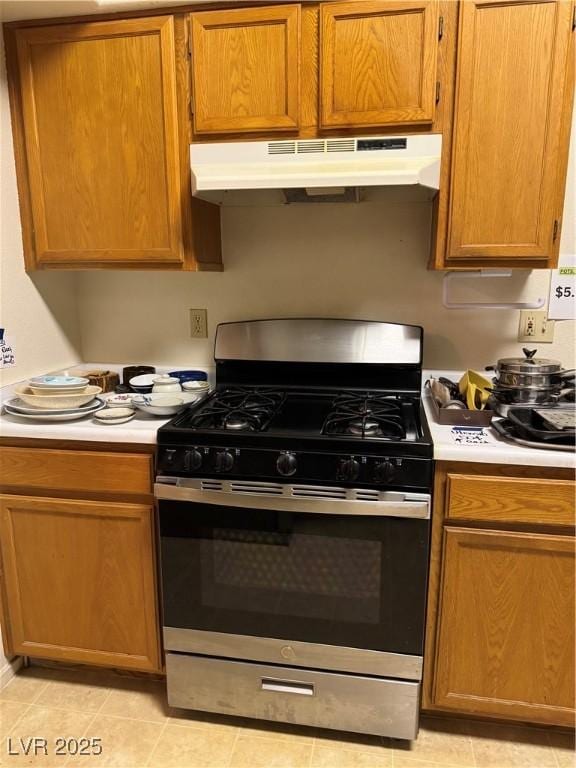 kitchen with stainless steel gas range oven, under cabinet range hood, light countertops, light tile patterned floors, and brown cabinetry