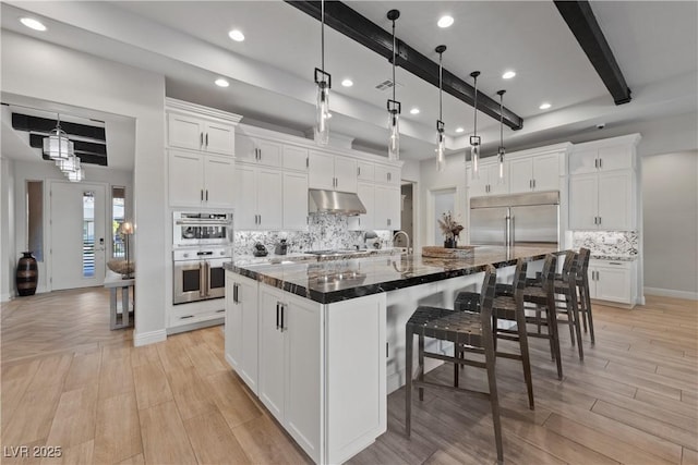 kitchen featuring white cabinets, appliances with stainless steel finishes, a breakfast bar, beam ceiling, and under cabinet range hood