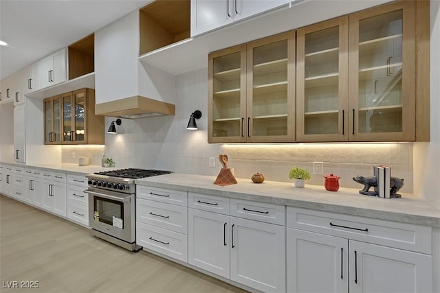 kitchen with stainless steel range, light wood-style flooring, decorative backsplash, custom range hood, and glass insert cabinets