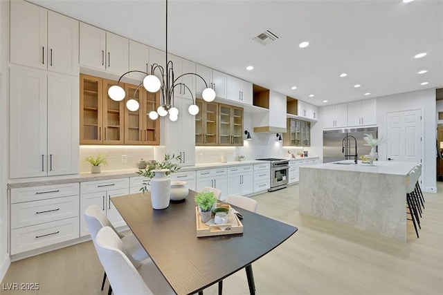 kitchen featuring visible vents, a kitchen island with sink, high end appliances, wall chimney range hood, and light wood finished floors