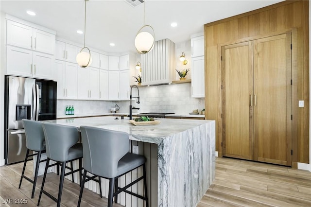 kitchen with decorative backsplash, a kitchen island with sink, white cabinets, stainless steel fridge with ice dispenser, and light wood-style flooring
