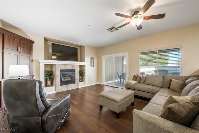 living room featuring ceiling fan, visible vents, dark wood finished floors, a tile fireplace, and baseboards