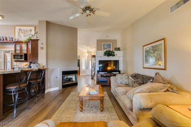living room featuring visible vents, wood finished floors, baseboards, ceiling fan, and a multi sided fireplace