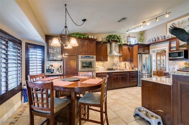 kitchen featuring wall chimney range hood, light stone countertops, hanging light fixtures, stainless steel appliances, and tasteful backsplash