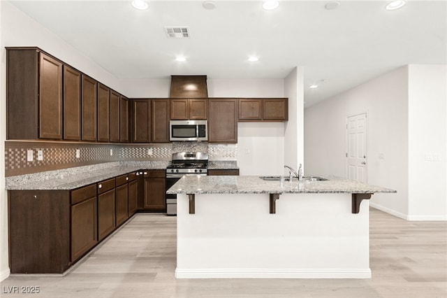 kitchen featuring a kitchen island with sink, light stone countertops, light wood-style flooring, backsplash, and stainless steel appliances