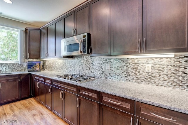 kitchen with dark brown cabinets, light stone countertops, appliances with stainless steel finishes, light wood finished floors, and backsplash