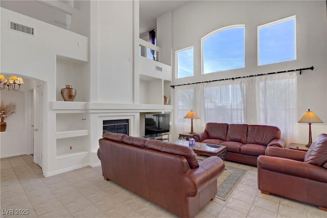 living area featuring an inviting chandelier, visible vents, a glass covered fireplace, a high ceiling, and built in shelves